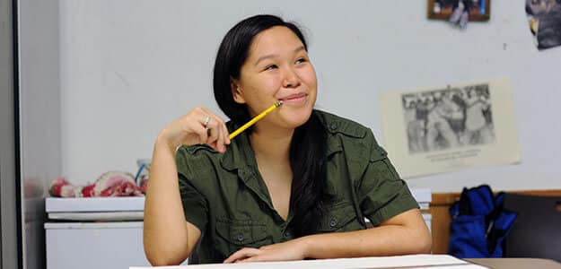 Young Alaska Native woman looks thoughtfully off toward something off-camera. She holds a extends the end of a pencil to her smiling lips.