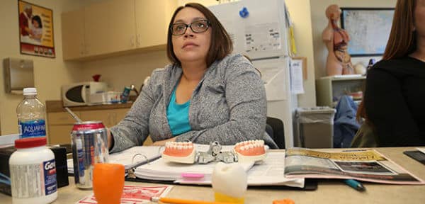 Alaska Native woman looks toward something off-camera while sitting in a medical-based classroom. She has notes and teeth model sitting in front of her.