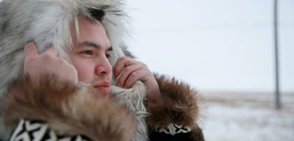 Young Alaska Native man holds up a parka hood around his face. He is outdoors.