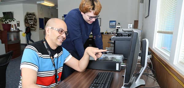 Two young men work in front of a desktop computer. One is pointing at the screen. The other is looking inside an open CPU.