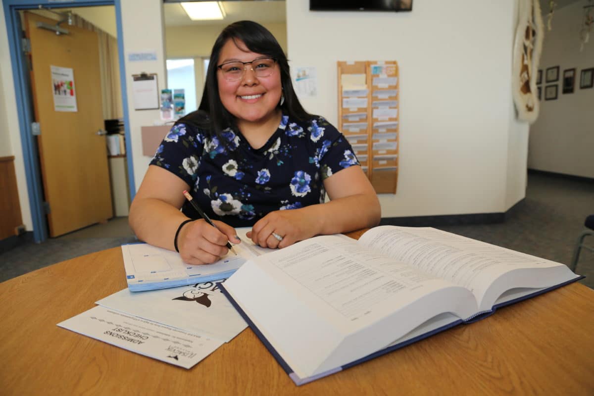 Smiling Alaska Native woman sits in front of open books and notes. She holds a pencil in her hand.